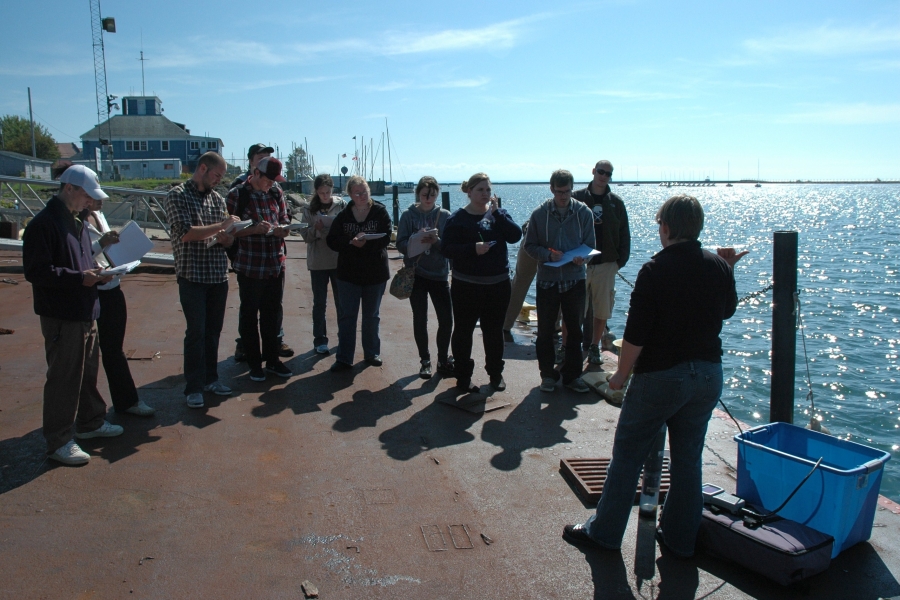 A group of people stand on a dock by the water taking notes. A person stands in front of them with some research equipment