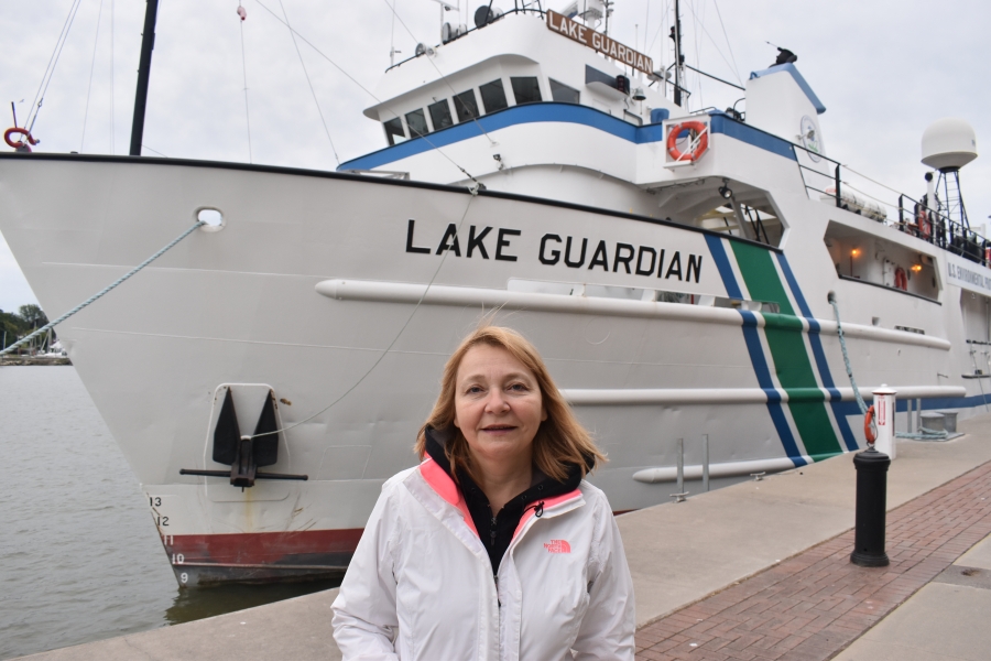 A person stands in front of the bow a large docked boat labeled "Lake Guardian."