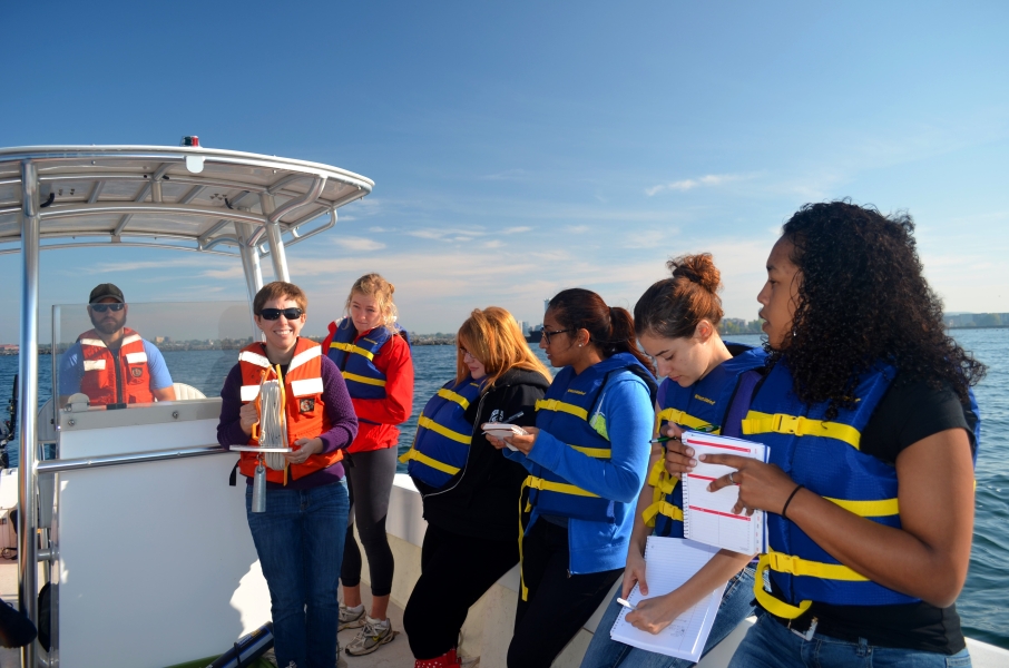 Several people wearing life jackets take notes on a boat while a person holds up a disk with a rope attached to it. Another person sits at the helm of the boat.