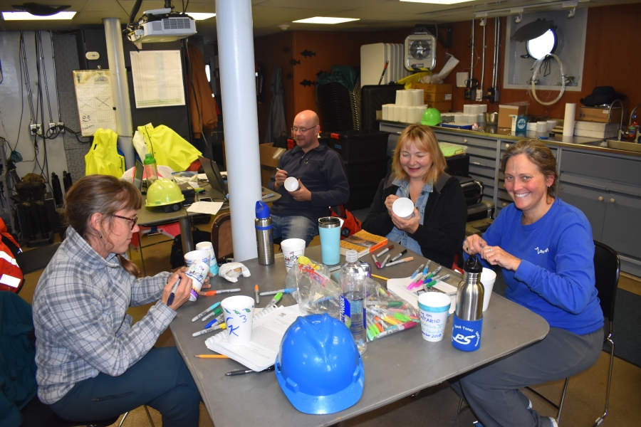 Four people sit in a lab. There are art supplies and they are drawing on styrofoam cups with markers. There is a hard hat on the table.