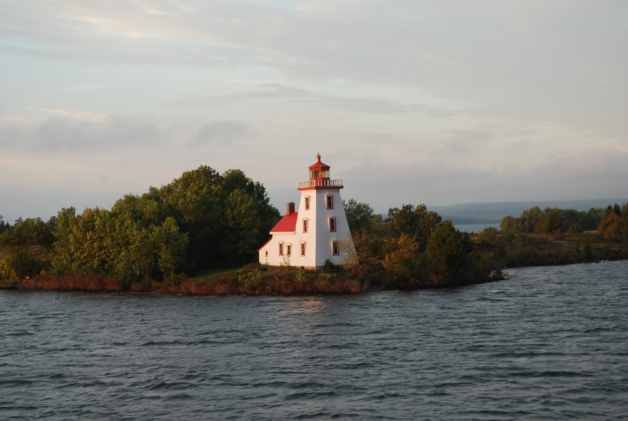 A lighthouse with a red roof on a wooded shoreline by the water.