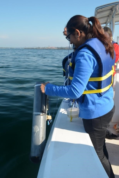 A different student stands by the edge of the boat, holding a plastic cylinder over the water.