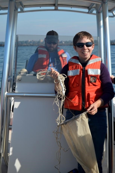 A person in a life jacket holds up a conical fabric net while another person stands at the controls of a boat. 