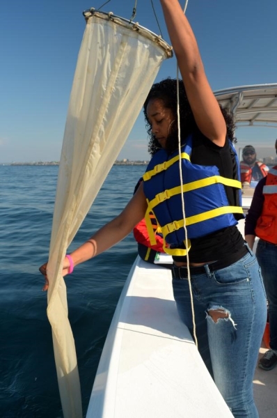 A student stands at the edge of a boat, pulling in a long fabric conical net.