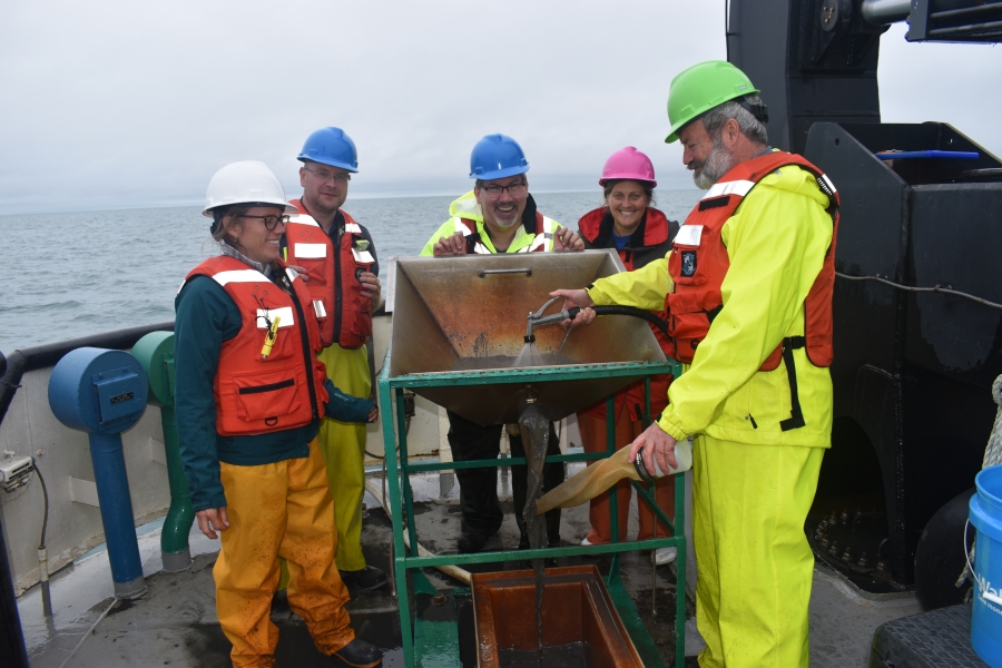 Five people stand around a metal basin on the back of a boat. One is spraying a hose into the basin.