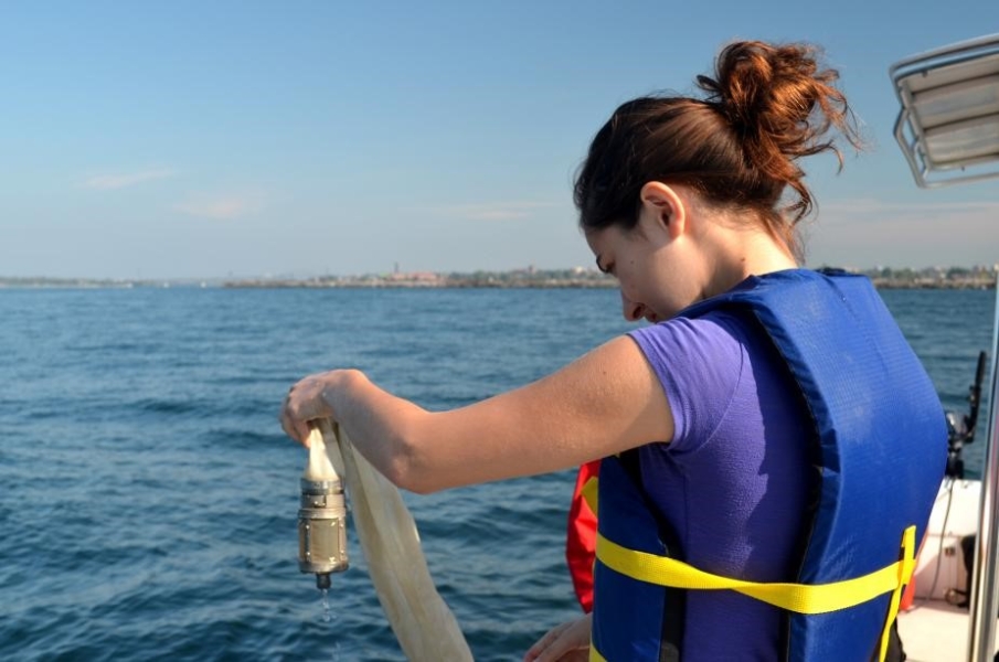 A student stands at the edge of a boat, holding up the little metal bucket at the end of a net.