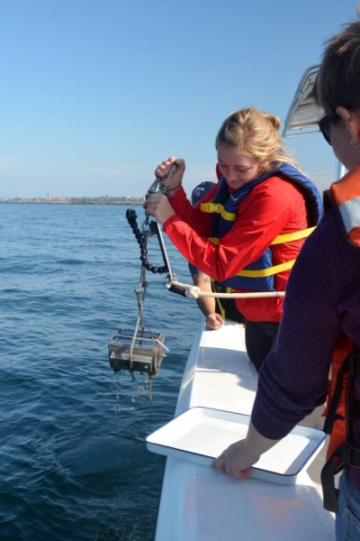 A student stands by the edge of the boat holding a metal grab sampler over the water