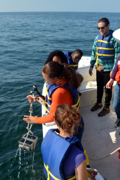 Students watch as one stands by the edge of the boat holding a metal grab sampler over the water