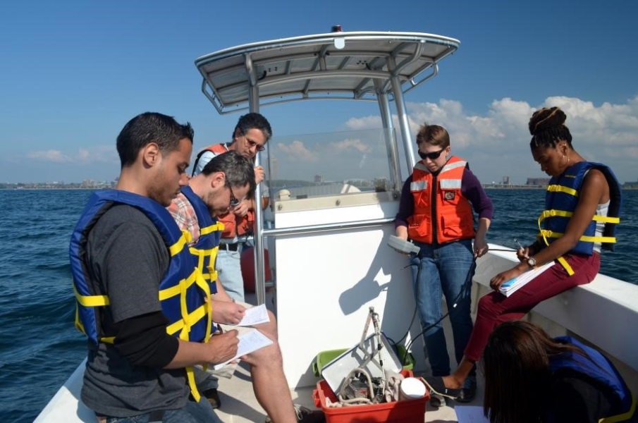 A class on a boat. A person looks at a instrument with a cable going into the water while other people take notes..