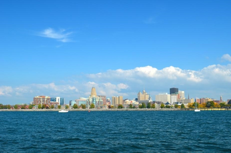 A clear sky  with a line of white clouds and water frame a city skyline. Two sailboats sit in the water