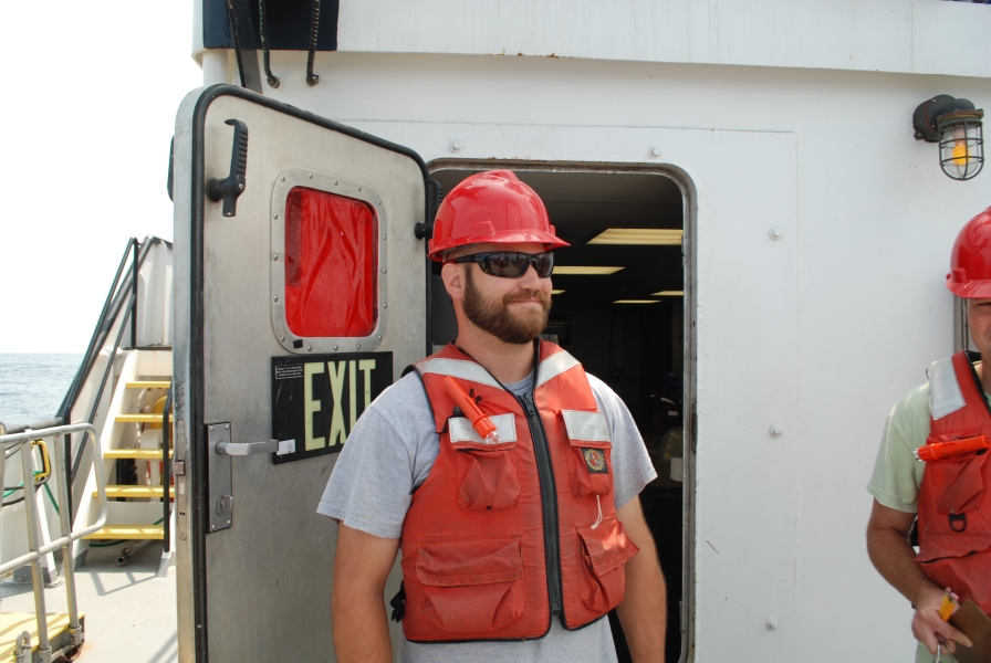 A person wearing a hard hat and life jacket stands by a bulkhead door labeled "exit" on a large boat