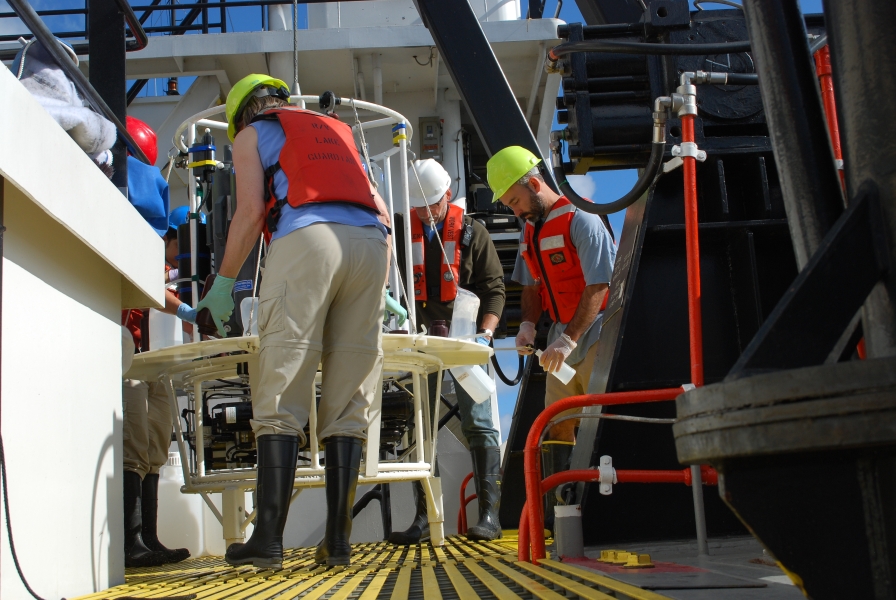 Several people in hard hats and life jackets fill bottles of water from a water sampler on the deck of a boat