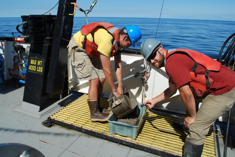 Two people in safety gear open a metal grab sampler into a tub and rinse the mud from the grab sampler into the tub while working on a large boat