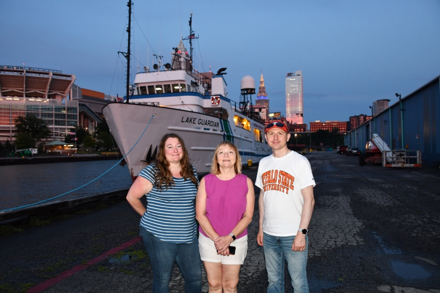 Three people standing in front of a ship docked in a city, a light-skinned ruddy woman with long brown hair and a striped blue and white shirt and jeans, a light-skinned blonde woman in a pink sleeveless blouse and white shorts, and a light-skinned man with a baseball cap and sunglasses and white Buffalo State University shirt and jeans