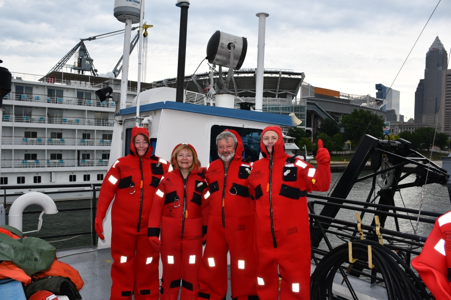 Four people wearing cold water safety suits pose on the deck of a ship in a city. One gives a thumbs up