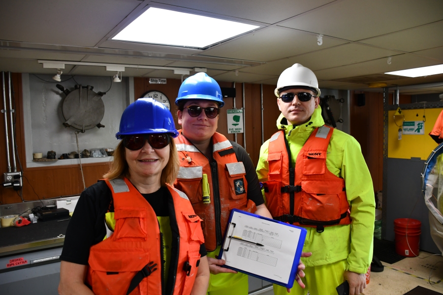 Three people wearing life jackets, hard hats, and sunglasses pose for a picture inside a boat. One is holding a clipboard with a data sheet and pencil