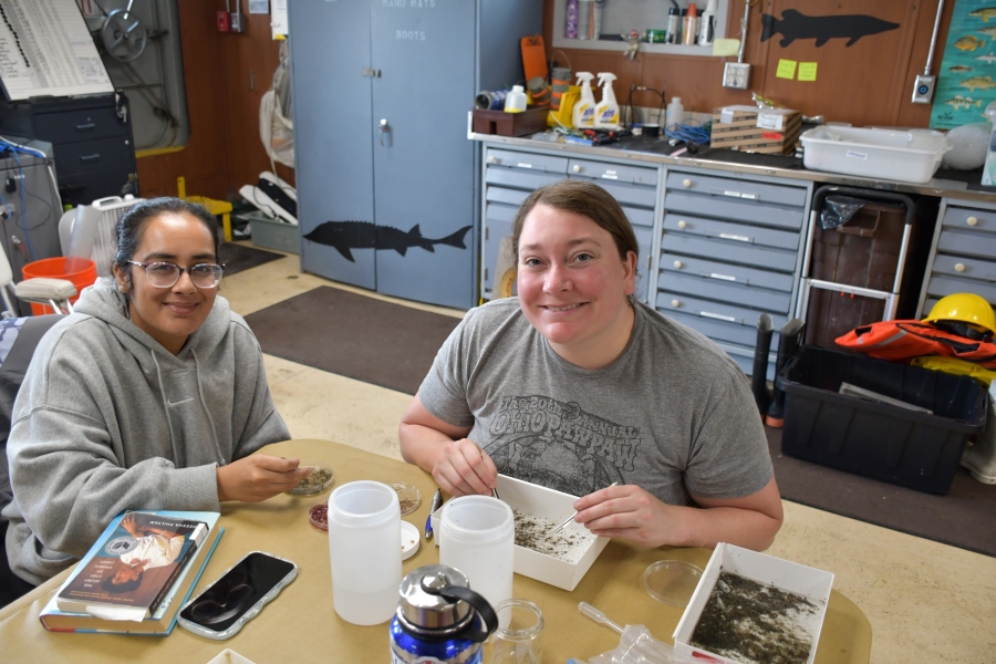 Two people pause and smile while sitting at a table in a lab on a boat and using forceps to pick organisms out of pans of sediment samples