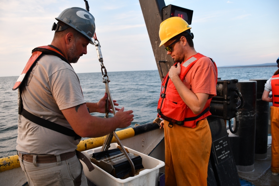 Two people in life jackets and hard hats working at the edge of a large boat. One person is holding a grab sampler in a tub preparing to open it