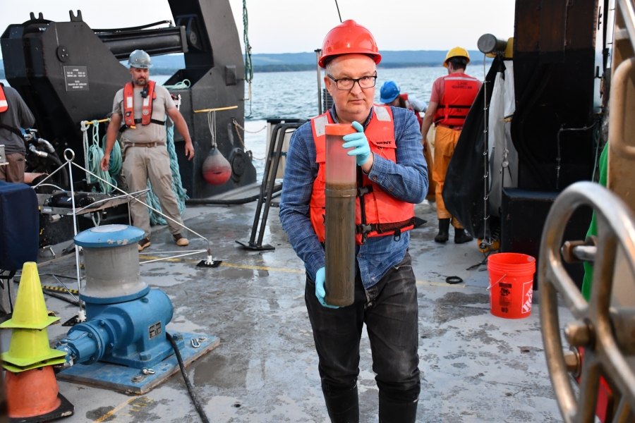 A person in a life jacket and hard hat carries a tube of a sediment core on the deck of a large boat as other people work in the background