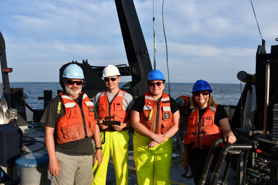 Four people in life jackets, hard hats, and sunglasses pose together on the deck of a large ship