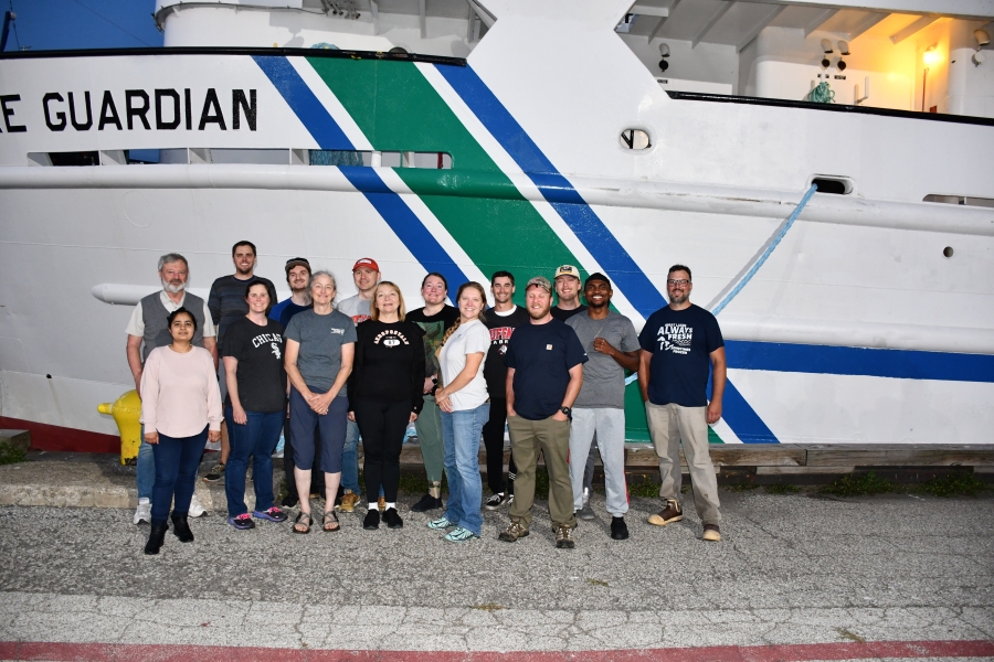 Fifteen people pose in two rose in front of a docked ship Lake Guardian