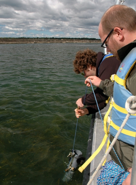 a student at the edge of the boat pulls up a plastic tube while another student helps handle the rope