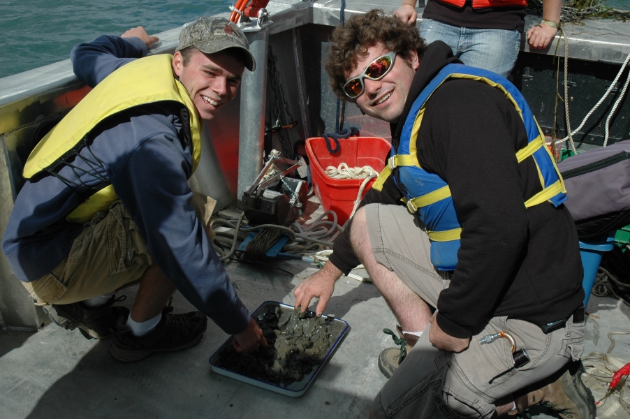 Two students wearing life jackets smile at the camera while kneeling down to touch the mud in a pan on the deck of a boat