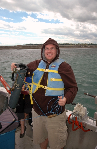 A person in a life jacket and hoodie stands on a boat holding up a water sampling bottle