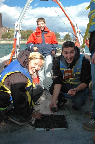 Two people kneel on the deck of a boat to play with mud in a pan on the deck. A person wearing a float jacket stands behind them. All three are smiling.