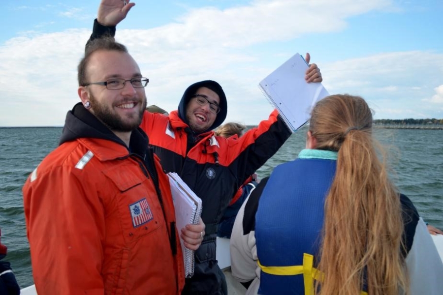 People on a boat. One is smiling at the camera, another has their arms in the air as they hold a notebook, and another has their back to the camera.