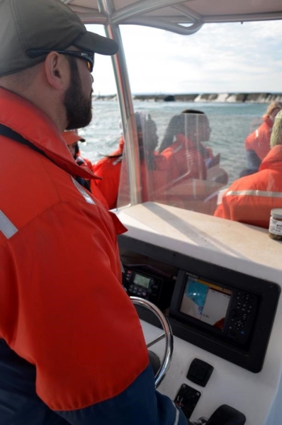 A person with a beard in cold water safety gear at the console of the boat.