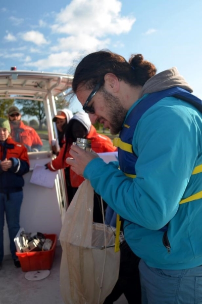 A person holding a net and a small metal cup. There are people on the boat in the background.