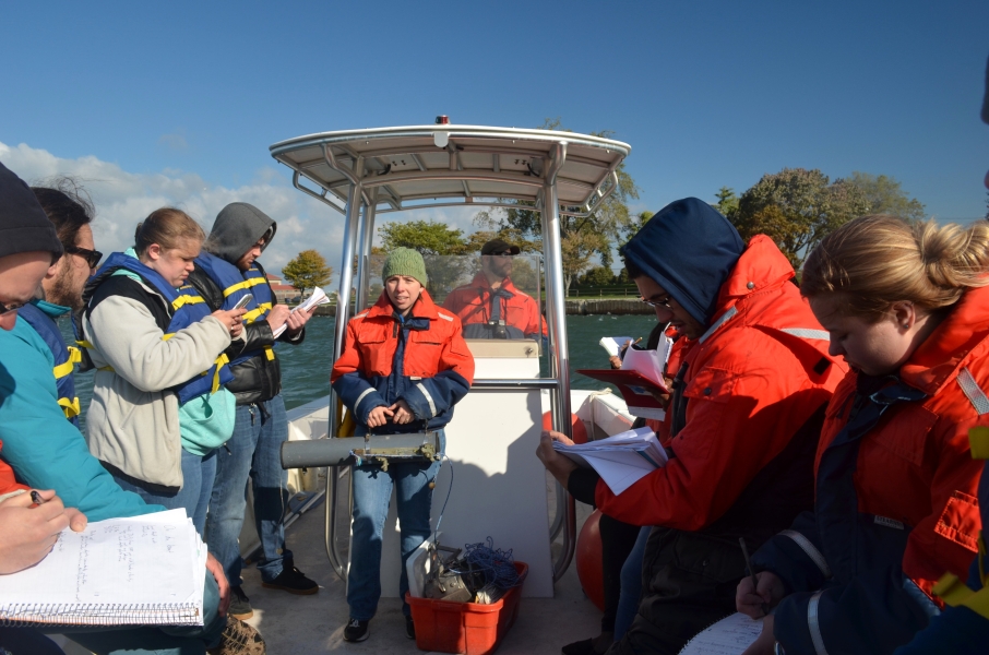 A person stands at the center of a boat holding up a cylindrical tube while other people take notes.