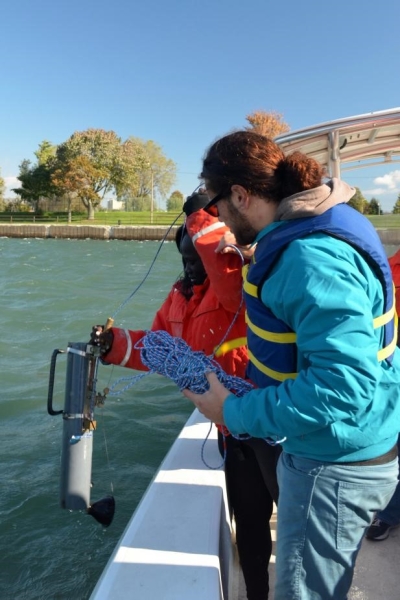 Two people stand at the edge of a boat. One person holds a cylindrical tube above the water while the other person holds the rope.