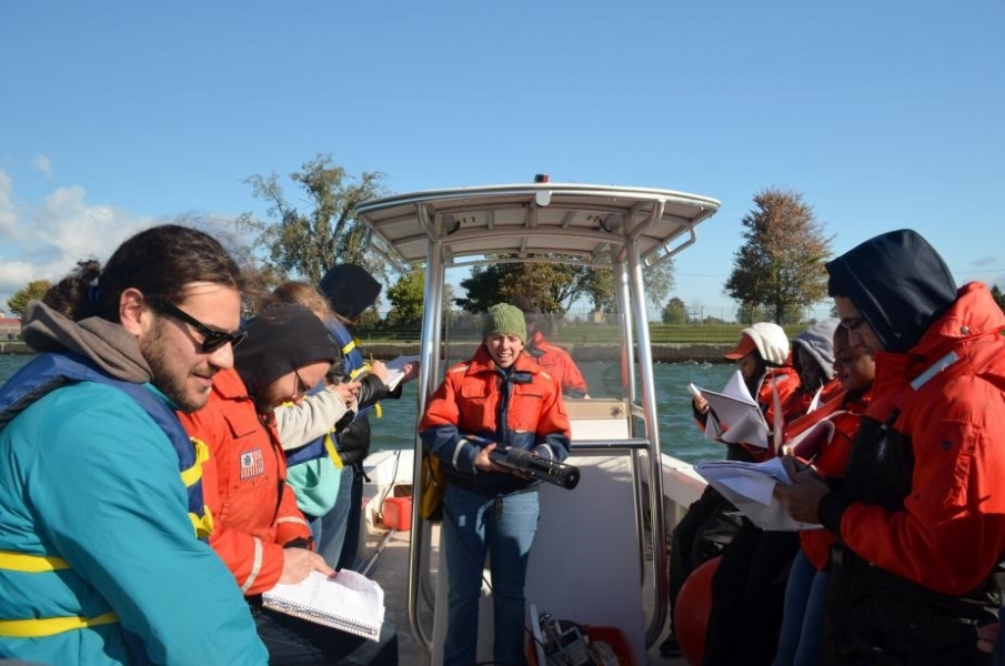 A person stands at the center of a boat holding up a cylindrical object while other people take notes.