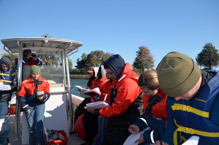 A person stands at the center of a boat holding up a cylindrical object while other people take notes.