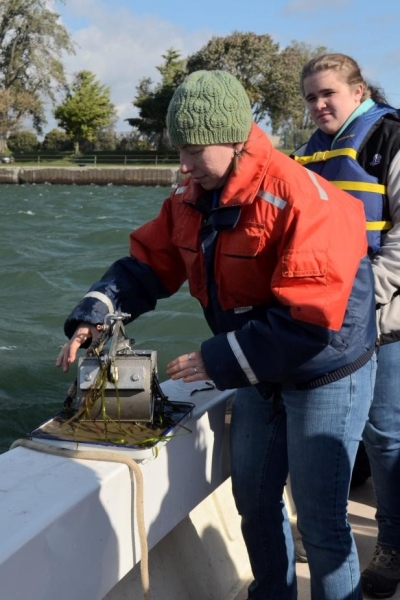 A person stands at the edge of a boat. They are opening a metal device that's on a pan, and there is mud and weeds coming out of the metal object.