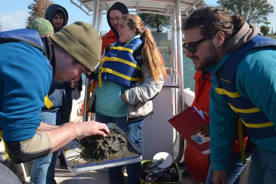 A person on a boat holds a pan with a big glob of mud and weeds on it. Other people are on the boat too.