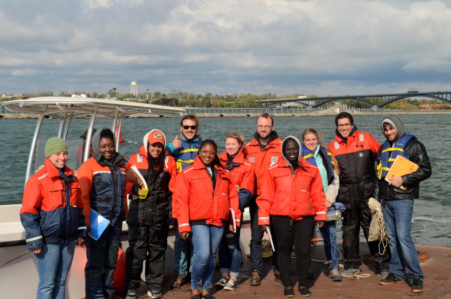 A group of ten people wearing cold water safety gear standing in front of a docked boat by the water.