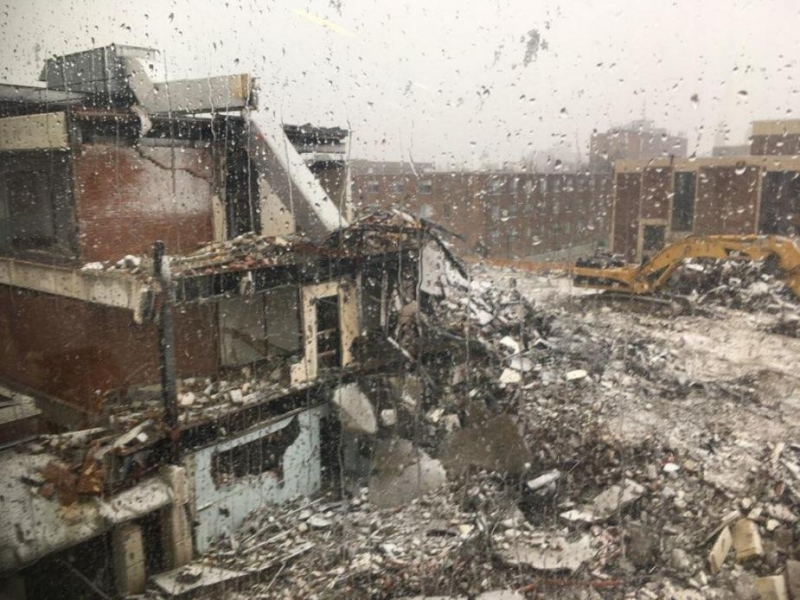 A construction site with a partially demolished three-story building with a light layer of snow covering the debris. The picture is taken through a window with water droplets on it.