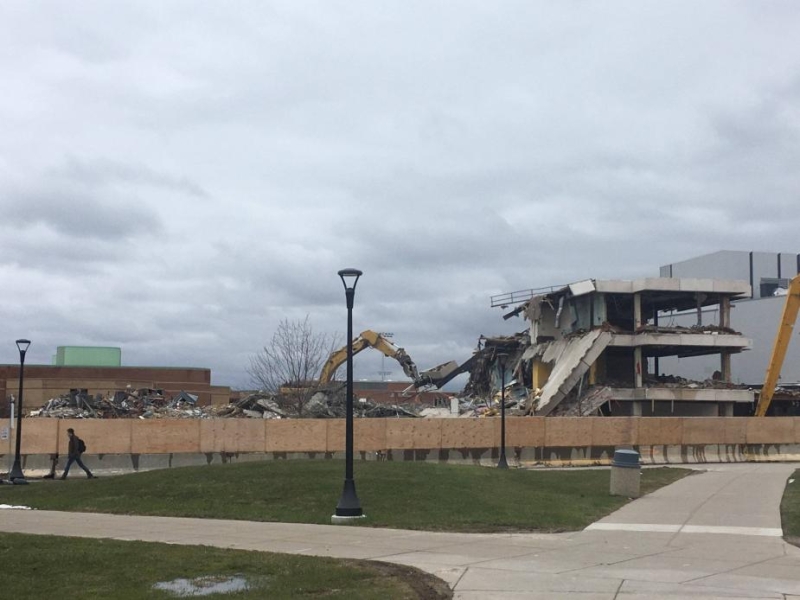 A construction site on a campus behind a wooden fence. An excavator is pulling a part off the second floor of a building. In the foreground are some sidewalks.