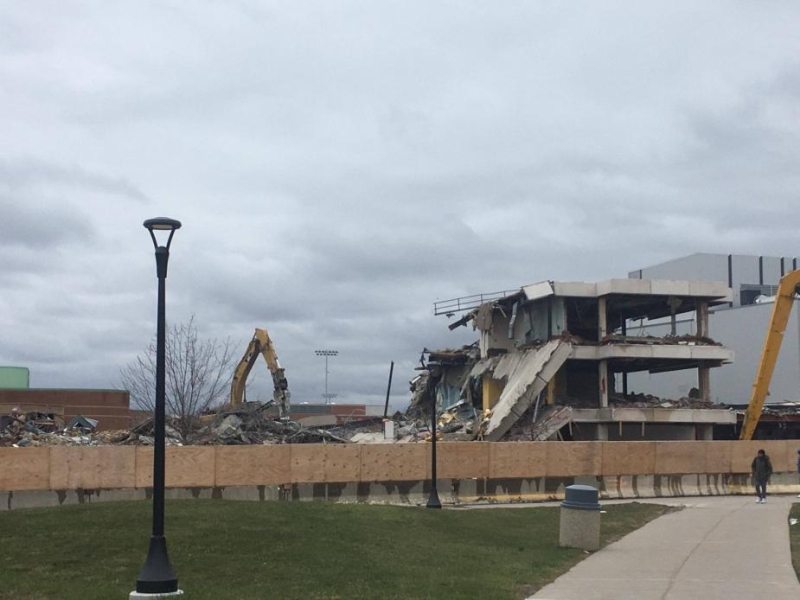 A construction site on a campus behind a wooden fence. An excavator is pulling a part off the second floor of a building. In the foreground are some sidewalks.