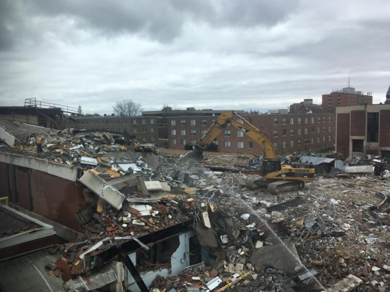 A construction site with a mostly demolished three-story building. Someone is spraying the building with water. The picture is taken through a window.