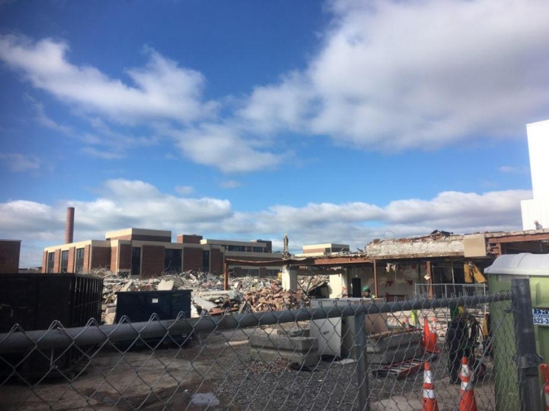 A view of a field of debris and a partially demolished building behind some construction equipment and a fence. The sky is blue with white clouds.