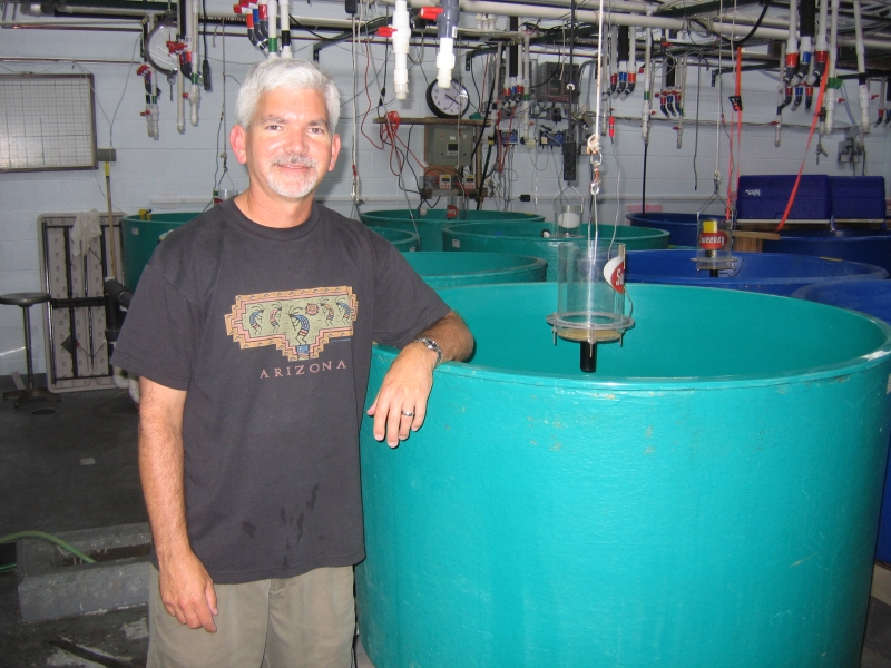 A person stands with their arm resting on a round green tank in a wet lab. Their shirt says "Arizona."