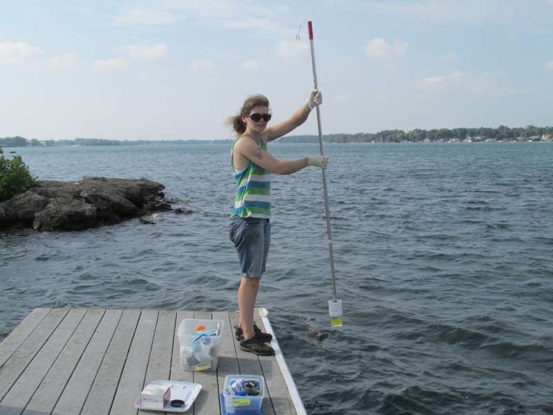 A graduate student stands at the edge of a dock, holding a pole with a cup at the end of it.