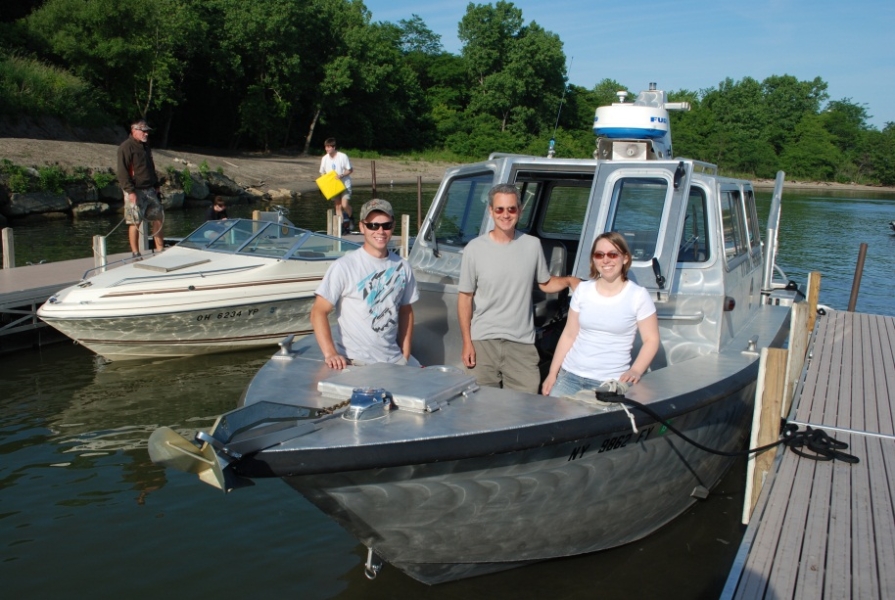 Three people stand for a picture at the front of a metal boat that's tied up to a dock. There are some other people launching a boat at the next slip over.