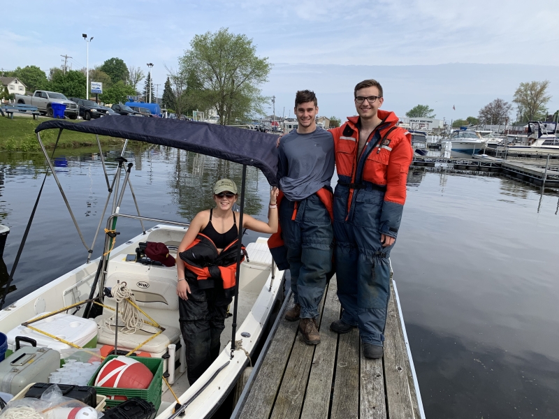 Two students stand on a dock near a boat with a bimini top while a third student stands in the boat next to them. They are all wearing full body flotation suits.