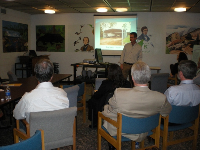 a group of people in chairs watch a presentation given by a different person at the front of the room. There is a presentation projected onto a screen, and murals painted on the walls of the room.