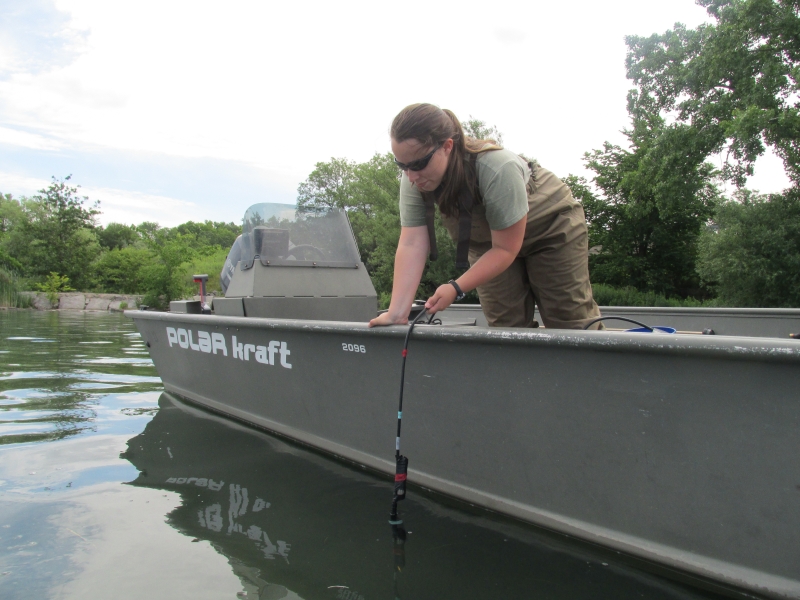 A graduate student leaning over the side of a boat to lower an instrument on a cable into the water.